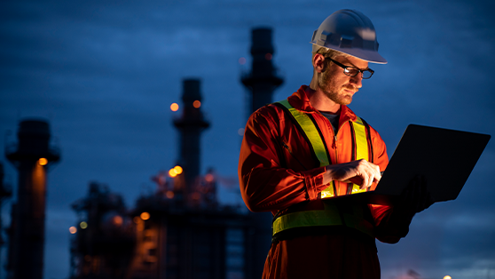 Man wearing hard hat looking at laptop standing in front of power station at night