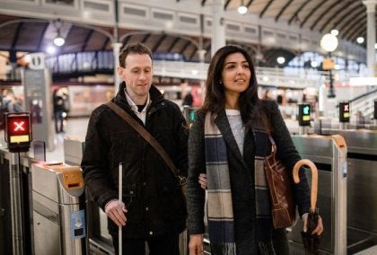 Woman and man using a white cane passing through ticket barriers in a station with arched roof