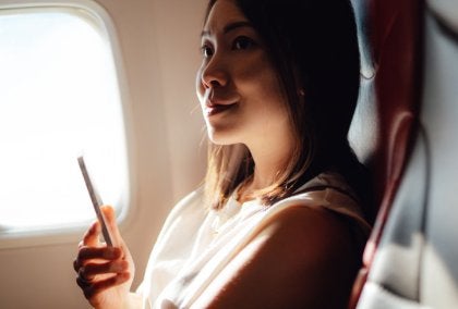 Woman in white tank top sitting in red airplane seat by window, holding smartphone