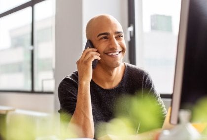 Person at desk talking on phone, computer monitor and potted plant nearby.