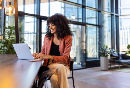 Smiling woman in blazer typing on laptop at a high table by floor-to-ceiling windows