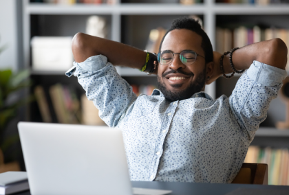 Smiling man with glasses leaning back at desk, laptop open, bookshelf behind him