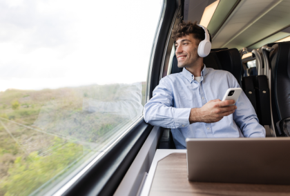 Young man with headphones holds phone and gazes out train window beside laptop