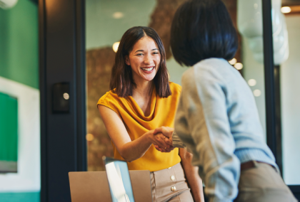 Two professional women smiling and shaking hands across a desk in an office.
