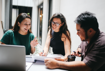 Three coworkers smiling and discussing papers by a laptop in an office.