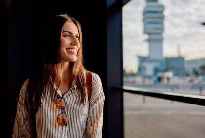Smiling young woman with sunglasses on shirt stands by window overlooking city tower