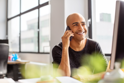 Smiling man at modern office desk talking on phone while looking at computer monitor