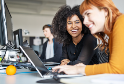 Photograph of two women smiling and collaborating on laptops in a bright modern office.