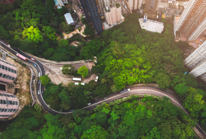 Aerial photograph of winding road and metro tracks through forested valley between skyscrapers.