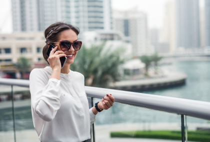 Smiling woman in sunglasses and white blouse on phone, holding car key fob by modern waterfront
