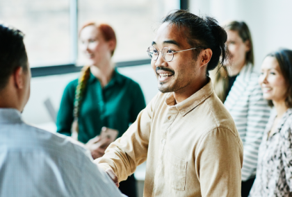 Man in tan sweater shaking hands with colleague as others smile behind