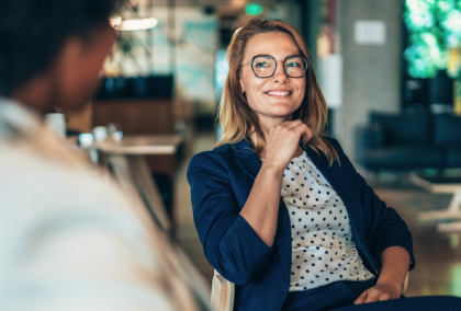 Photograph of a smiling woman in glasses and blazer resting her chin on her hand in a modern office