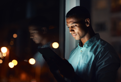 Young man in teal shirt reading a tablet by a window at night with blurred city lights