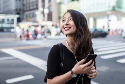 Photograph: smiling young woman wearing earphones and holding a smartphone on busy city crosswalk