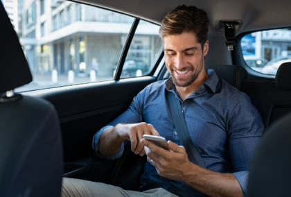 Photograph of a man smiling in a car backseat, using his phone with city view outside.