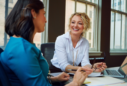 Two businesswomen at an office desk smiling and reviewing documents beside a laptop.