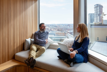 Two colleagues chatting on a cushioned window bench in modern office overlooking city skyline.