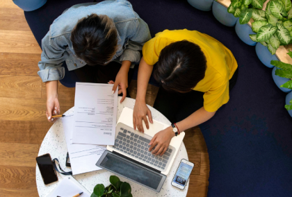 Overhead photo of two people at a round table, one reviewing papers and one using a laptop