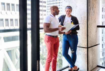 Two coworkers chat by floor-to-ceiling windows in a bright office, holding papers and coffee