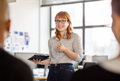 Smiling red-haired woman with glasses holds tablet and gestures in office presentation