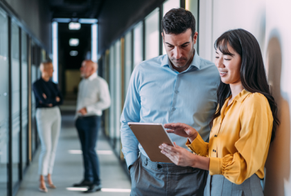 Two office colleagues lean on a hallway wall reviewing a tablet, with coworkers walking behind.