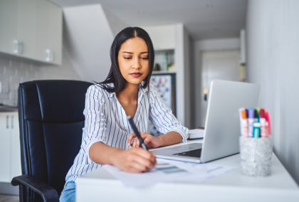 Woman seated at desk in home office, writing notes beside open laptop