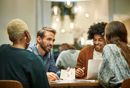 Photo of four diverse coworkers chatting around a laptop at a cafe-style office table