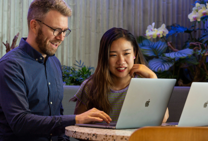 Two colleagues at a wooden desk smiling as they collaborate on laptops