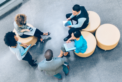 Five colleagues seated on circular ottomans in a modern lobby, collaborating with laptops