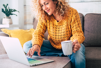 Curly-haired woman in yellow sweater on sofa typing on laptop while holding coffee mug