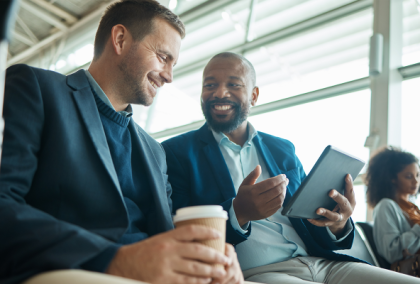Two men in suits smiling on a plane, one with coffee and the other with a tablet.