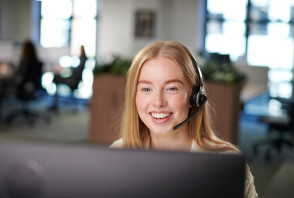 Photograph of a smiling woman wearing a headset at an office desk, blurred colleagues behind