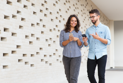 Man and woman walking by a white perforated wall, smiling as they check smartphones.