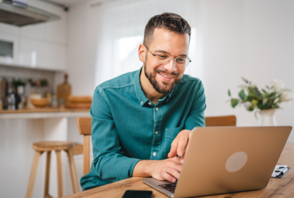 Smiling bearded man in green shirt and glasses typing on laptop in bright kitchen