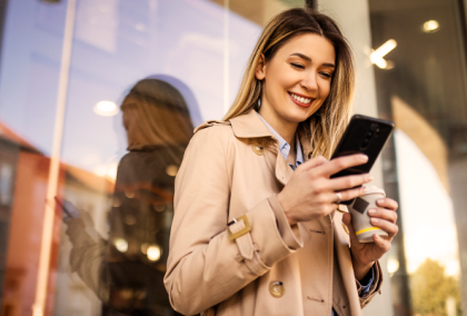 Smiling woman in trench coat using smartphone outside glass storefront