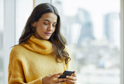 woman in yellow sweater looking at smartphone by window with city skyline in background