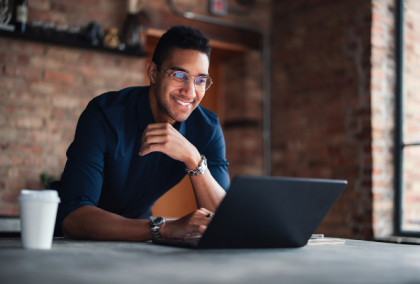 Photograph of a smiling man with glasses working on a laptop at a desk against a brick wall