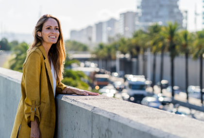 Young woman in mustard coat leaning on a barrier overlooking busy street with palm trees.