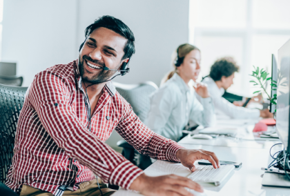 Smiling headset-wearing customer service agent typing at desk with colleagues blurred in background