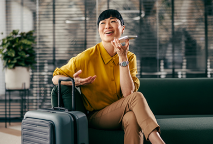 Passenger wearing mustard shirt sits in airport lounge checking phone, silver carry-on beside.