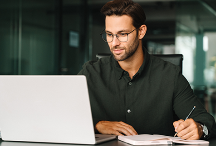 Photo of a man writing notes beside an open laptop in a modern office setting.