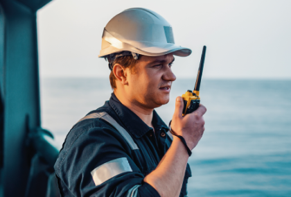 Ship officer in white hard hat and coveralls speaking into a handheld radio on deck