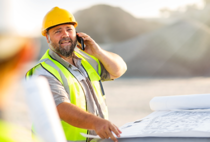 Smiling bearded construction worker in hard hat and vest checks blueprints and talks on phone