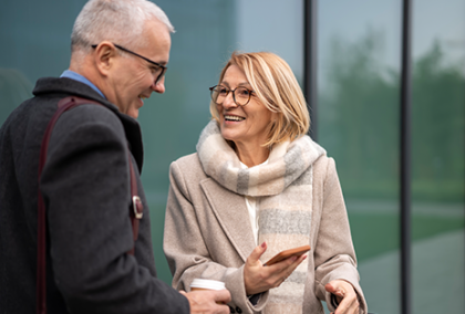 Man in dark coat holds thermos, talks with woman in white coat holding phone outside glass building