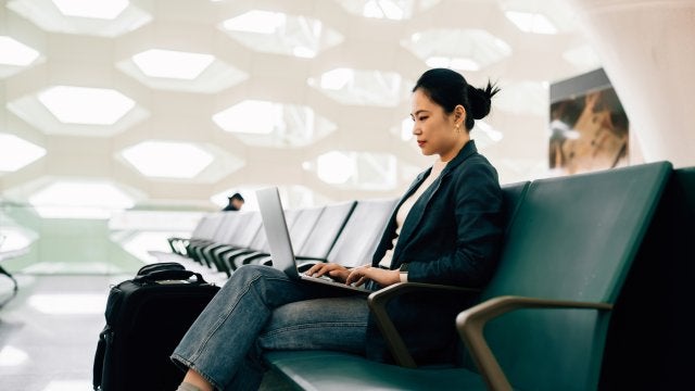 Profesional trabajando en una laptop desde la sala de espera de un aeropuerto mientras gestiona actividades relacionadas con viajes corporativos.