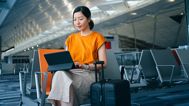 A woman in an orange shirt sits at an airport, working on a laptop beside her suitcase.