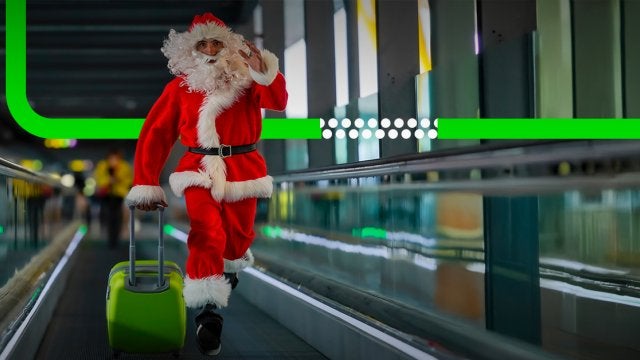 Persona disfrazada de Santa Claus caminando apresurado en un aeropuerto moderno mientras arrastra una maleta verde, transmitiendo humor y caos navideño.