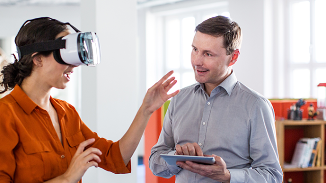 Lady with a VR headset on, with a man in a blue shirt talking to her