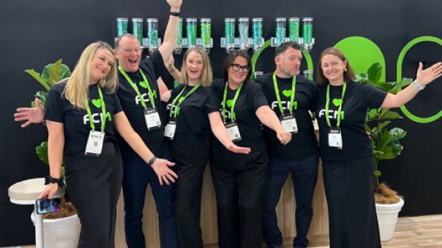 Seven women in black shirts and green lanyards smiling with arms outstretched by green leaves