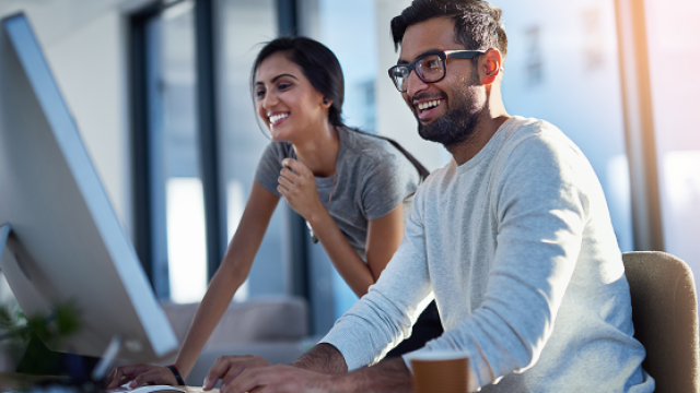 Photograph of two smiling coworkers at a desktop computer in a modern office